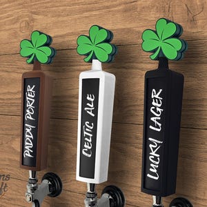 May include: Three beer tap handles with shamrock toppers. The handles are brown, white, and black, each with a different beer name: "Paddy Porter", "Celtic Ale", and "Lucky Lager". The handles are mounted on a wooden background.