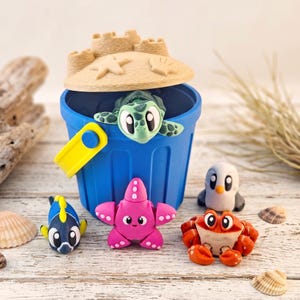 May include: A blue plastic bucket with a yellow handle and a sandcastle-shaped lid. Inside the bucket is a green sea turtle figure. Outside, there are four small animal figures: a blue and yellow fish, a pink starfish, a white penguin, and a red crab.