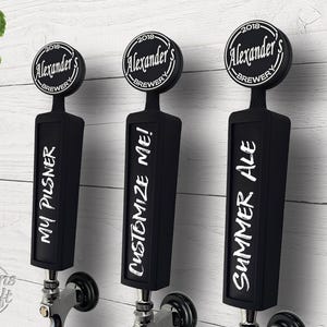 May include: Three black beer tap handles with white text. The handles read "My Pilsner", "Customize Me!", and "Summer Ale". Each handle has a circular top with "Alexander's Brewery 2018" printed on it. The background is a white wooden wall.