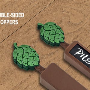 May include: Two double-sided beer tap toppers with green hop designs. One topper has the word "Pilsner" in white. The text "DOUBLE-SIDED TOPPERS" is in white. The toppers are brown and green.