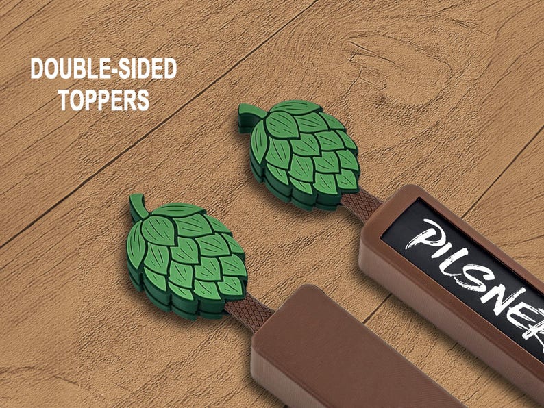 May include: Two double-sided beer tap toppers with green hop designs. One topper has the word "Pilsner" in white. The text "DOUBLE-SIDED TOPPERS" is in white. The toppers are brown and green.