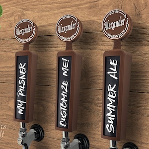 Op de afbeelding: Drie bruine biertapkranen met zwarte schoolbordlabels. De handgrepen zijn gelabeld met "My Pilsner", "Customize Me!" en "Summer Ale" in witte letters. Elke handgreep heeft een cirkelvormige bovenkant met "Alexander's Brewery" erop gedrukt. De achtergrond is een houten muur.