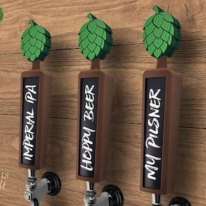 May include: Three beer tap handles with green hop-shaped tops and chalkboard-style labels. The labels read "IMPERIAL IPA", "HOPPY BEER", and "MY PILSNER". The handles are mounted on a wooden wall.