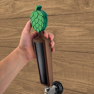 May include: A brown and black beer tap handle with a green hop design. The tap handle is held against a wooden background.