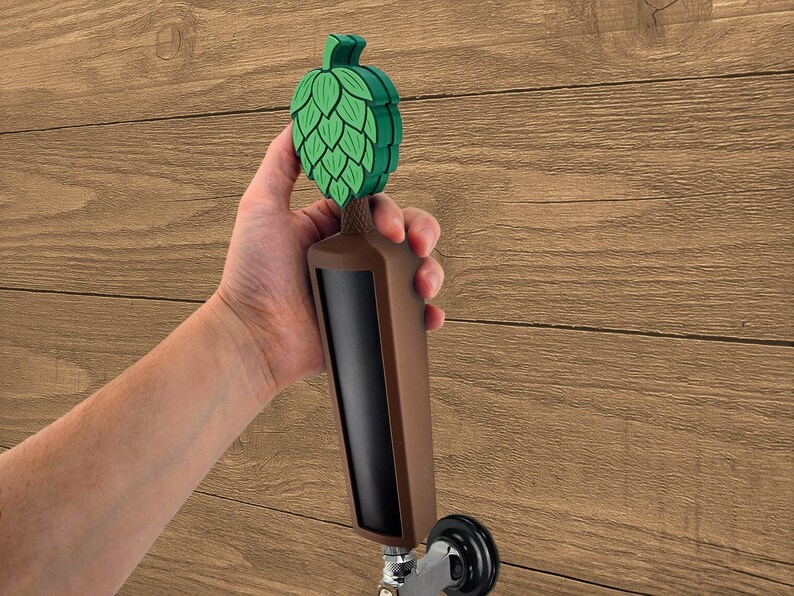 May include: A brown and black beer tap handle with a green hop design. The tap handle is held against a wooden background.