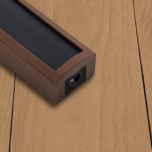 May include: A rectangular, brown object with a black top and a black end. The object has a metallic appearance and is set against a light brown wooden surface. The object has a hole on the end.