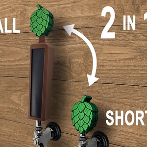 May include: A brown and black tap handle with a green hop design. The image shows two tap handles, one tall and one short, with the text "2 in 1". The word "TALL" is above the tall handle, and "SHORT" is below the short handle.
