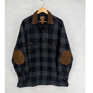 Peut inclure: Une chemise à manches longues boutonnée avec un motif à carreaux gris foncé et noir. Le vêtement présente un col en velours côtelé marron et des coudières marron assorties sur chaque manche. Deux poches poitrine boutonnées sont visibles.