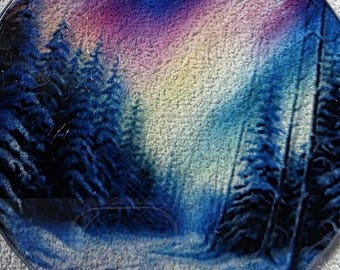 May include: Round Christmas ornament featuring a winter forest scene with a path leading through snow-covered trees. The sky transitions from blue to purple and yellow. A red ribbon is attached for hanging.