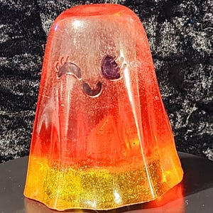 Può includere: Una statuetta di fantasma in resina rossa e oro con un viso che fa l'occhiolino e dettagli glitterati.
