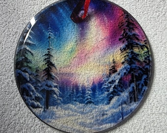 May include: Round Christmas ornament featuring a winter forest scene with snow-covered trees under a colorful aurora borealis. The ornament has a red ribbon for hanging. The artwork is a vibrant mix of blues, purples, and yellows.