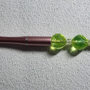 Puede incluir: Un bolígrafo malva con punta negra y extremo decorativo. El extremo del bolígrafo presenta dos grandes cuentas facetadas de color verde lima y cuentas transparentes más pequeñas. Es un instrumento de escritura.