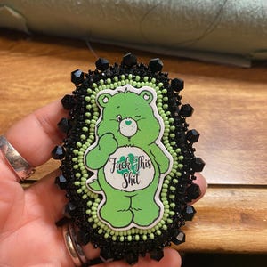 May include: A green Care Bear-style pin with the text "Fuck This Shit". The pin is surrounded by black and green beads and black crystal accents. The pin is an oval shape.