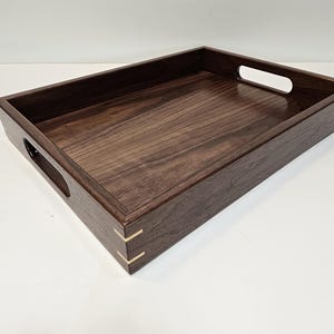 May include: A rectangular wooden serving tray with a dark brown finish. The tray has cut-out handles on the short sides and brass accents at the corners. The wood grain is visible on the tray's surface.