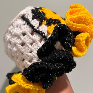 以下が含まれることがあります： 白、黄色、黒のグラニー・スクエアのデザインが施された手作りのかぎ針編みの帽子。 黄色と黒のフリル付きつば、銀色のメタリック糸のアクセントが特徴です。 ユニークでカラフルなアクセサリーです。