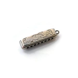 May include: A small, rectangular silver-toned metal harmonica charm featuring an engraved eagle design on the top surface. The instrument has a series of rectangular air holes along the side and a small circular loop at one end for attachment.
