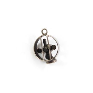 May include: A silver-toned metal charm shaped like a small desk fan. The object features a circular outer frame with two vertical bars protecting a central three-bladed propeller. A small circular jump ring is attached to the top for hanging.