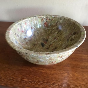 May include: A speckled, round bowl with a wide rim. The bowl has a mix of colors, including green, brown, red, and black. The bowl is sitting on a wooden surface.