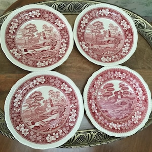 May include: Four round ceramic plates featuring a red and white transferware pattern. Each plate displays a detailed landscape scene with a stone bridge, trees, and buildings, surrounded by a decorative floral border along the scalloped edges.