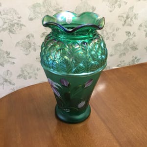 May include: A green glass vase with a ruffled rim and leaf pattern. The vase has a flared base and is decorated with painted purple and white flowers. The vase is sitting on a wooden surface.