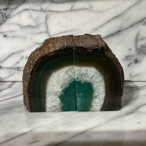 May include: A pair of bookends crafted from a single sliced agate geode. The exterior features a rough, brown, natural stone crust, while the interior reveals concentric bands of dark green and white crystalline quartz with a sparkling center.