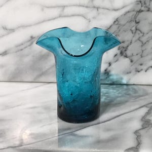 May include: A vibrant turquoise blue glass vase featuring a distinct crackle texture throughout the body and a wide, ruffled, undulating rim. The glass transitions to a slightly deeper shade of blue at the solid, flat circular base.