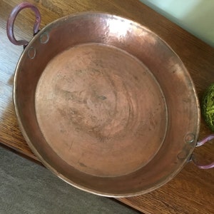 May include: A hammered copper pan with two curved handles. The pan has a wide, shallow bowl and a reddish-brown color. The handles are a darker shade of red. The pan is likely used for cooking.
