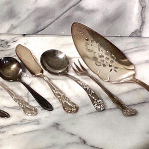 May include: A collection of antique silver serving utensils, including a fork, spoon, butter knife, ladle, and cake server. The cake server has a decorative floral design. The utensils are arranged on a marble surface.
