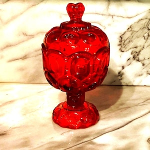 May include: A ruby red glass candy dish with a heart-shaped finial on the lid. The dish has a round body with a repeating pattern and sits on a pedestal base. The glass is transparent, allowing light to pass through.