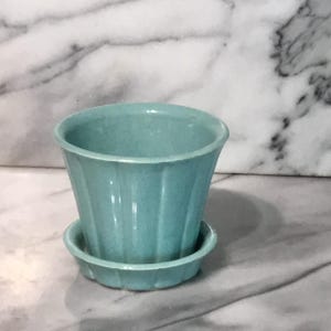 May include: A small, turquoise ceramic flower pot with a matching saucer. The pot has a fluted design and a slightly flared rim. The saucer is round with a similar fluted edge. The pot and saucer are sitting on a marble surface.