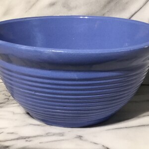 May include: A periwinkle blue ceramic mixing bowl. The bowl has a wide, rounded shape with a slightly flared rim. Horizontal ridges encircle the exterior, adding texture. The bowl is likely for kitchen use.