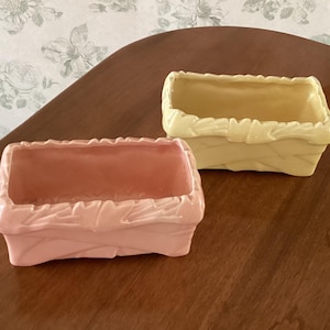 May include: Two rectangular ceramic planters. One is pale pink, the other is light yellow. Both have a textured, woven-like design around the edges. The planters are empty and sit on a dark wooden surface.