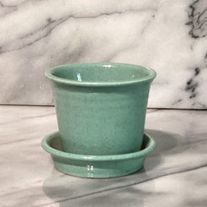 May include: A small, turquoise ceramic planter with a matching saucer. The planter has a slightly flared rim and a speckled finish. The saucer is round with a raised edge. The set is designed for small plants and adds a touch of color.