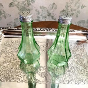 May include: Two vintage green glass salt and pepper shakers with silver-colored metal tops. The shakers have a geometric design and are displayed on a silver tray with ornate detailing. The background features a floral wallpaper.