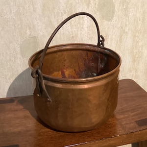 May include: A round copper pot with a hammered texture and a dark, weathered patina. The vessel features a sturdy, arched metal handle attached to two side brackets, with visible signs of oxidation and wear on the interior and exterior surfaces.