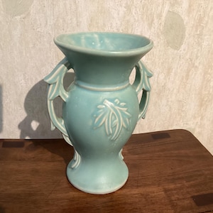 May include: A light blue ceramic vase with a wide flared rim and two decorative handles on the sides. The body of the vase features a raised leaf-like pattern on the front and back, with a smooth, glossy finish and a pedestal base.