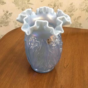May include: A light blue, iridescent glass vase with a ruffled rim. The vase has a floral pattern and a label. The vase is sitting on a wooden surface.