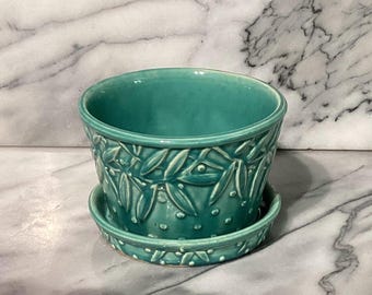 May include: A teal ceramic planter with a matching saucer. The planter features a raised leaf and dot pattern around the exterior. The planter is round with a smooth interior and sits on a matching saucer, perfect for indoor plants.