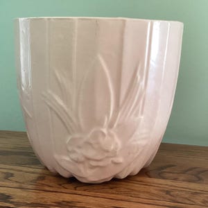 May include: A light pink ceramic planter with a floral design. The planter has vertical ribbed detailing and a sculpted flower and leaf motif. The planter is sitting on a wooden surface.