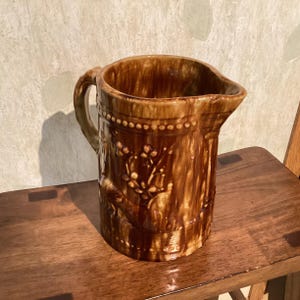 May include: A brown ceramic pitcher with a handle and a spout. The pitcher has a textured surface and is decorated with a floral design. The pitcher is sitting on a wooden surface.