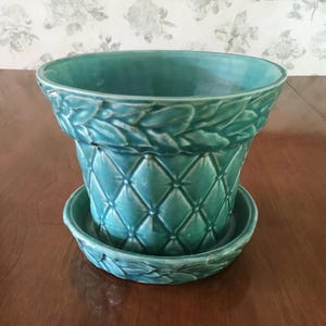May include: A teal ceramic plant pot with a matching saucer. The pot features a raised diamond lattice pattern on the body and a decorative braided or leaf-like relief design around the upper rim and the edge of the circular base saucer.