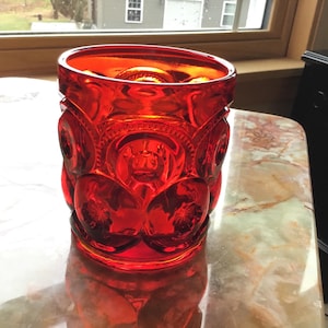 May include: A vibrant red glass tumbler with a detailed, embossed pattern. The design features repeating starburst and floral motifs. The glass is thick and appears to be vintage, with a rich, saturated color. The tumbler is sitting on a marble-like surface.