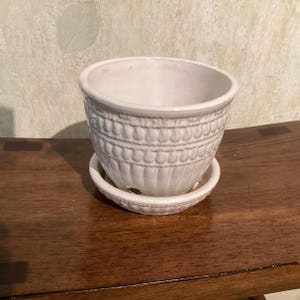 May include: A white ceramic plant pot with a matching saucer. The exterior of the pot features a textured pattern of vertical ridges and a horizontal band of embossed teardrop shapes around the upper section, with a smooth interior finish.
