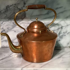 May include: A polished copper tea kettle featuring a wide, rounded body, a curved spout, and a domed lid. The kettle has a tall, arched brass handle with a dark brown wooden grip centered at the top, providing a functional and classic appearance.