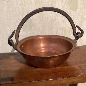 May include: A round copper bowl with a smooth, polished interior and a wide rim. The bowl features a dark, textured metal swing handle attached to two curved, snake-shaped metal mounts on opposite sides of the exterior rim.