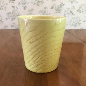 May include: A light yellow ceramic vase with a speckled texture and a subtle, wavy pattern. The vase has a cylindrical shape and is displayed on a wooden surface. The background features a floral wallpaper.