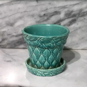 May include: A teal ceramic plant pot with a matching saucer. The pot features a raised diamond-quilted pattern on the body and a decorative leaf-motif border around the top rim and the edge of the saucer, finished with a glossy glaze.