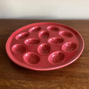 May include: A round, red ceramic escargot plate with 12 individual wells. The plate has a slightly raised rim and is designed to hold escargot. The plate is a solid, vibrant red color.