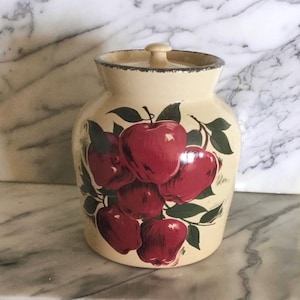 May include: Cream-colored ceramic jar with a lid, decorated with a cluster of red apples and green leaves. The jar has a slightly flared shape and a dark rim. A small knob sits atop the lid.