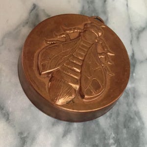 May include: A round, copper-colored object with a raised relief of a bee. The bee is detailed with wings, legs, and body segments. The object has a circular shape and a slightly raised edge.